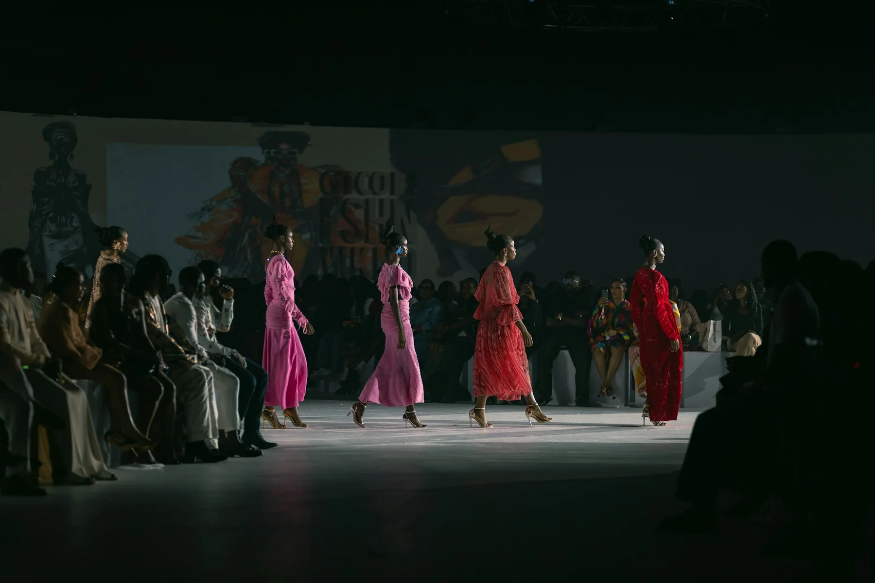 Models in bold color walk a darkened runway past a seated audience